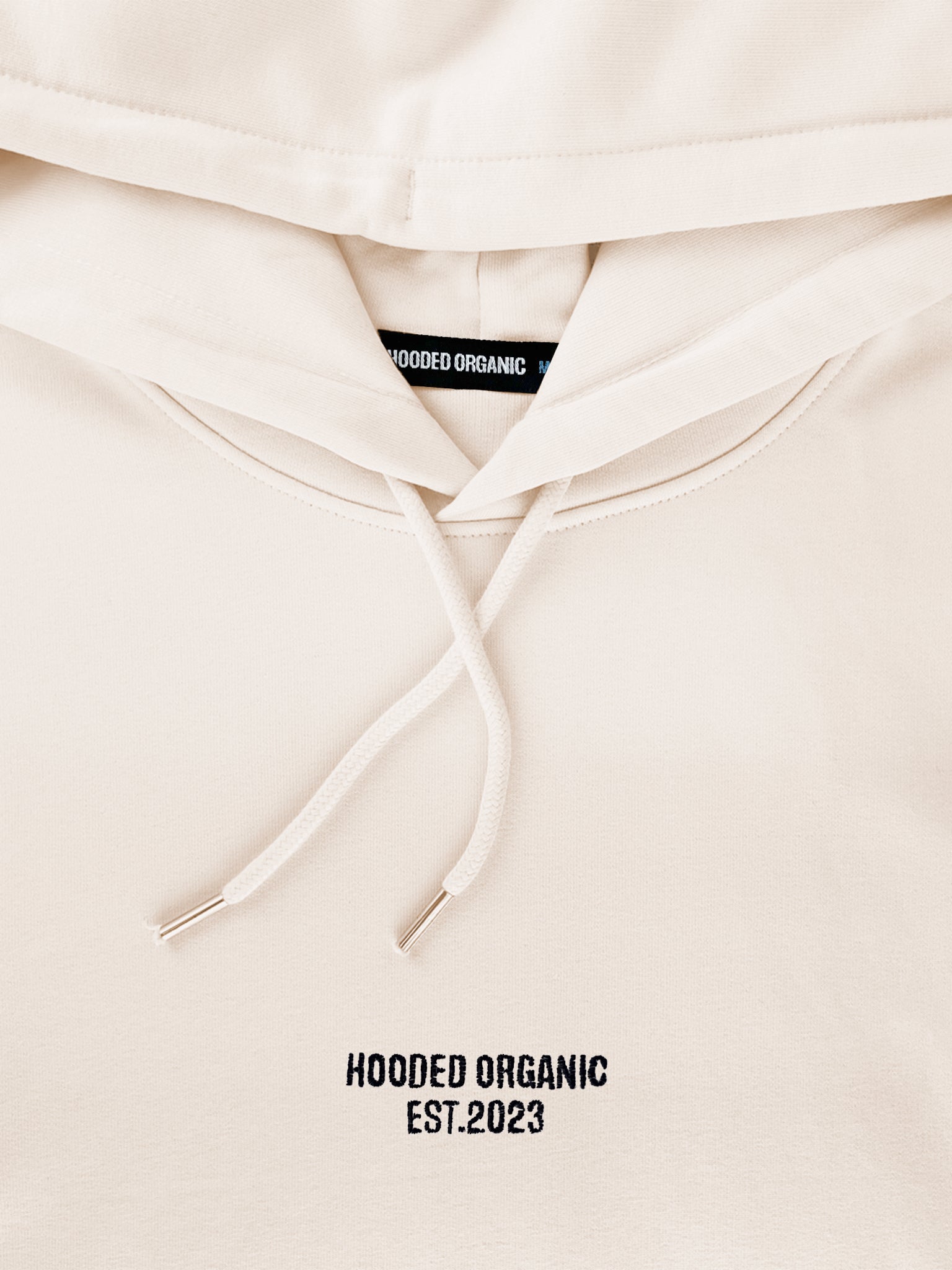 Nærbillede af Hooded Organic-logo på beige hoodie – lavet i 100% økologisk bomuld
